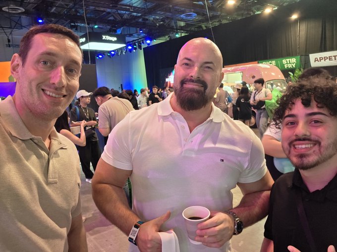 Three men are smiling at the camera at what appears to be a gaming convention, with signs for 'X-Stag', 'STIR FRY', and 'APPET' visible in the background.