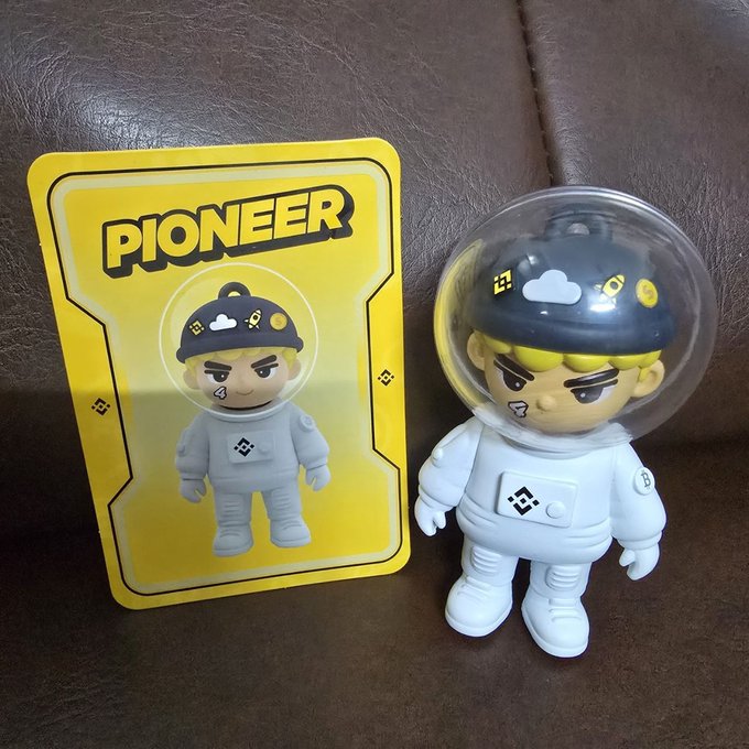 An astronaut figurine wearing a spacesuit and helmet sits next to a yellow card labeled "PIONEER," matching the celebratory post about a Binance gift that embodies the spirit of Web3 exploration.