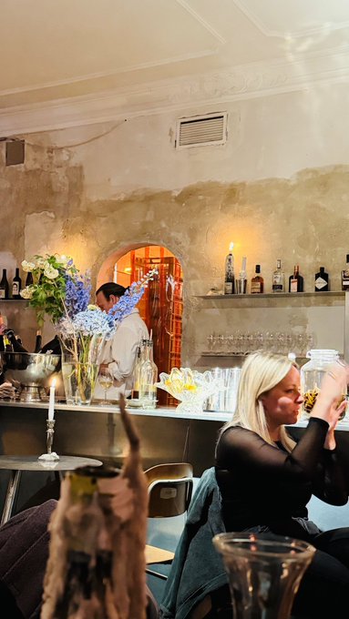 A warmly lit bar scene with a blonde woman in the foreground, a bartender pouring drinks behind the counter, fresh flowers, and an eclectic, artsy interior.