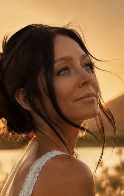 A portrait photograph of a woman with long dark hair blowing in the wind, looking over her shoulder with a slight smile against a warm golden sunset background.