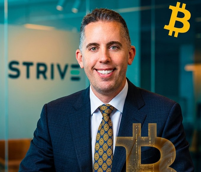 The image features a man dressed in a suit, smiling at the camera while sitting behind a desk with a computer monitor. The background of the photo shows a glass window that displays the word "Strive" written in white letters. There are also two other words visible on the window - "bitcoin" and "bear market proof". These words seem to be related to the Bitcoin project, as indicated by the post's title.