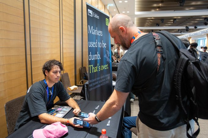 A man with a backpack looks at a smartphone at a table, while a seated man behind a banner reading "Markets used to be dumb. That's over." for Fables watches.