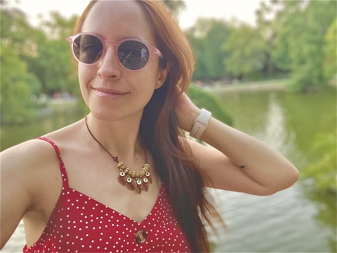 A woman in pink sunglasses and a polka dot top poses by a serene lake. The "Gm fam" caption pairs with her relaxed, cheerful morning vibe in the sunlit park setting.