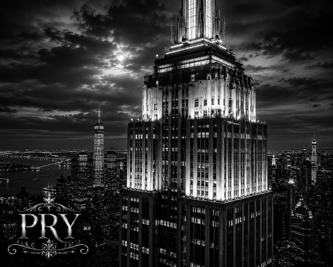 A black-and-white aerial nighttime view of the Empire State Building in New York City, illuminated against a cloudy sky with the One World Trade Center visible in the distance; the watermark 'PRY' appears in the lower left.