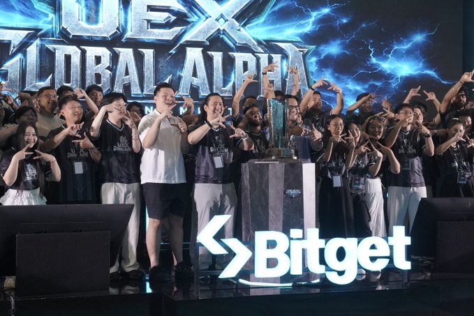 A group of excited participants pose together at a Bitget-hosted UEX Global Alpha Tournament event, with many flashing "shaka" hand signs in front of a trophy on a branded stage. The "Bitget" logo and "UEX Global Alpha" branding are prominently displayed, tying the celebratory photo to the kickoff of the global alpha trading competition.