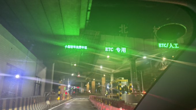 A nighttime view from inside a vehicle approaching a highway toll plaza illuminated in green, with signs indicating lanes for license plate recognition (小型车不免车牌), ETC, and manual (人工) payment.