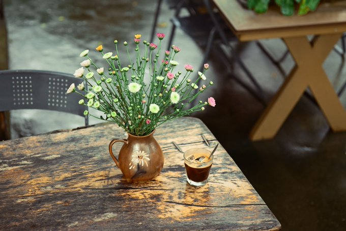 A rustic wooden table holding a small brown ceramic pitcher filled with colorful wildflowers (pink, yellow, and white) alongside a glass of dark beverage with straws.