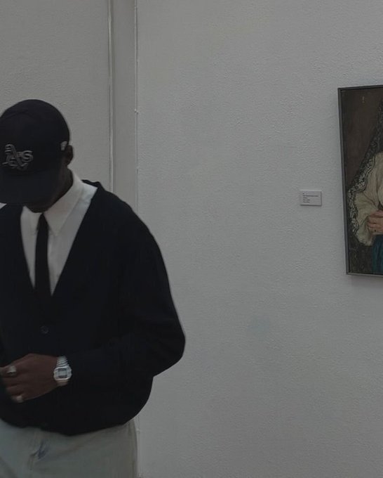A person wearing a dark 'LA' baseball cap, white collared shirt, dark cardigan, and wristwatch stands against a white wall near a partially visible framed painting.