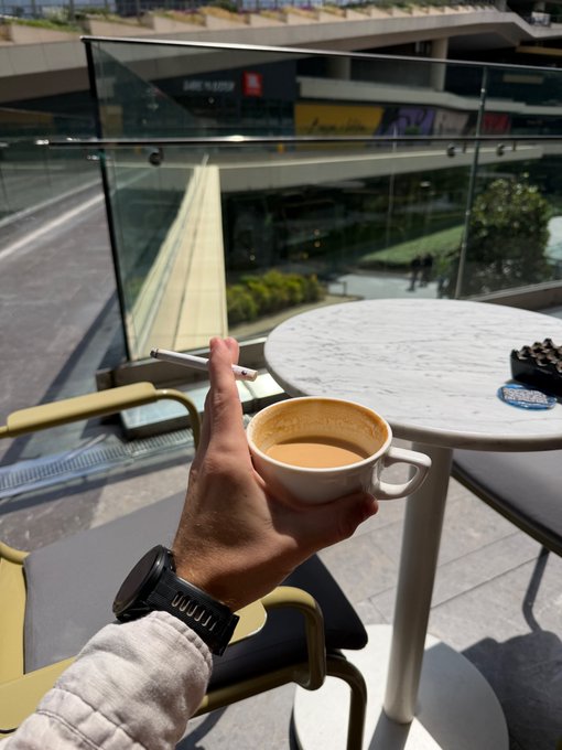 The image shows a person holding a cup of coffee, possibly enjoying their time outdoors during the summer season. They are seated at an outdoor table with chairs, which appears to be located near a glass wall or railing. The scene suggests that they might be taking a break from work or simply relaxing in a comfortable setting while sipping on their coffee and watching people pass by outside.