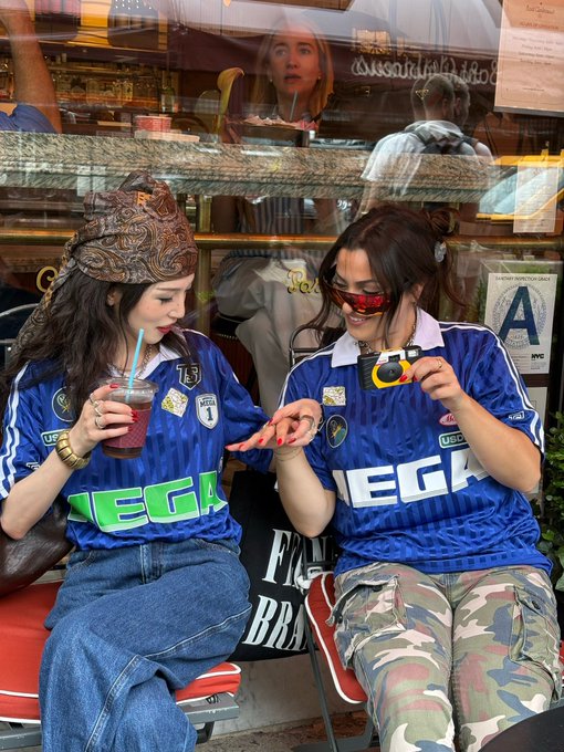 Two young women sit outside a bakery wearing matching blue MEGA jerseys, with one showing off her red-painted nails while holding a purple smoothie and the other flashing a peace sign in sunglasses and camo pants—rocking the @megaeth MOSS jersey mentioned in the post, complete with a "#1" patch and "MEGA" branding across the chest.