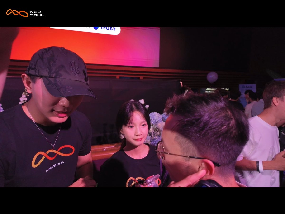 A man in a black cap and black NeoSoul t-shirt stands next to a young girl at a dimly lit event with red and purple lighting, while another person wearing glasses faces them in the foreground.