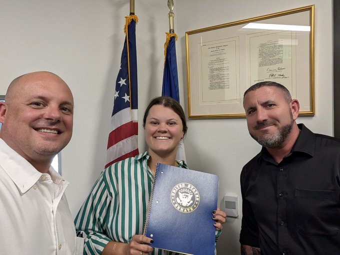 Three people pose smiling in front of an American flag and a framed document, with a woman in a striped shirt holding a blue "Colorado Department of Regulatory Agencies" binder. The image captures the in-person advocacy effort referenced in the post, showing two supporters alongside Colorado State Chapter President Derek Porter delivering regulatory clarity messaging to Senator Hick's field office.