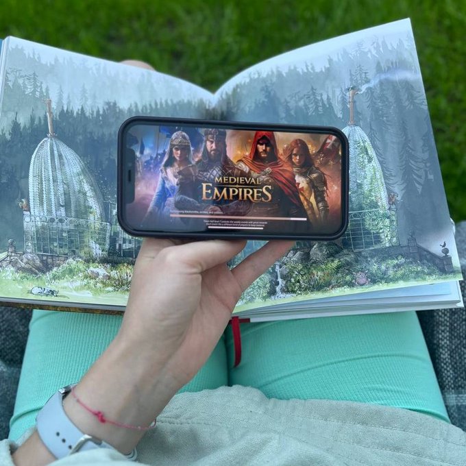 A person holding a smartphone displaying the loading screen for 'Medieval Empires' over an open illustrated book resting on their lap outdoors.