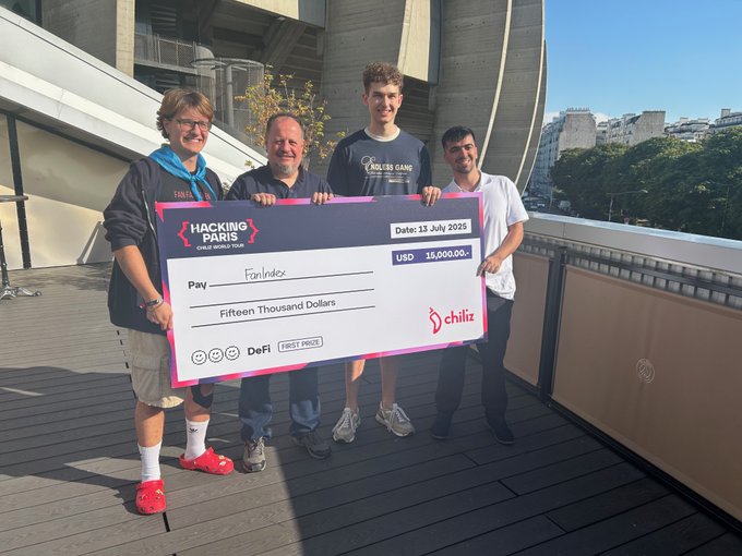 Four men pose outdoors on a wooden deck holding a large novelty check from "Hacking Paris" made out to "FanIndex" for "Fifteen Thousand Dollars" dated 13 July 2025, featuring the "chiliz" logo.