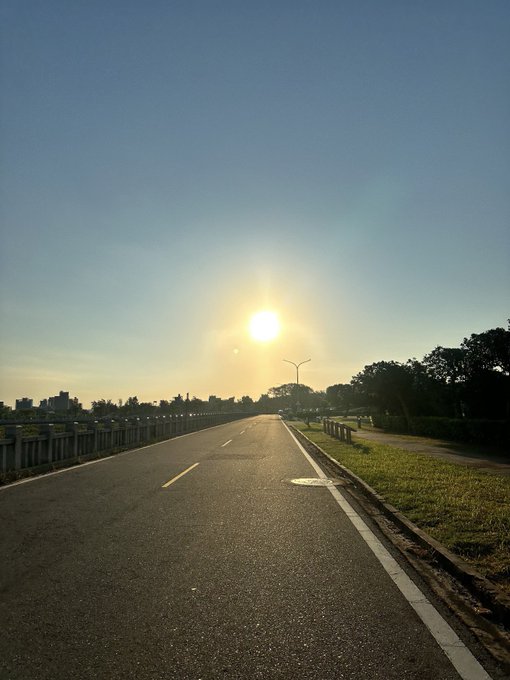 A photo taken from a road perspective showing a bright sunset or sunrise over a highway with streetlights, trees, and a clear sky.