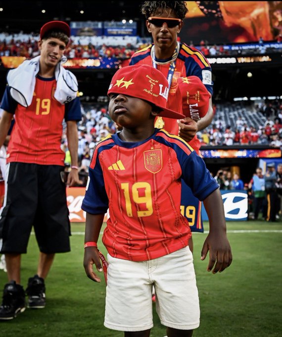 A young child wearing a red Spain national team jersey with the number 19 and a championship hat stands on a soccer field with players behind him.