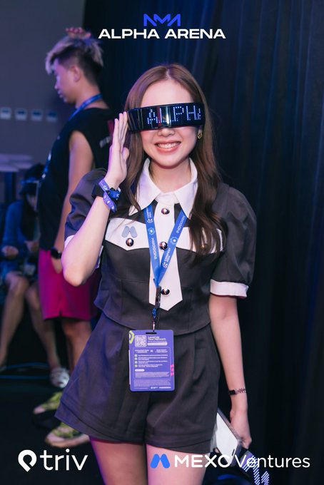 A photograph from the 'Alpha Arena' event (branded with Triv and MEXC Ventures logos) shows a smiling woman in a maid outfit wearing LED glasses that display scrolling text.
