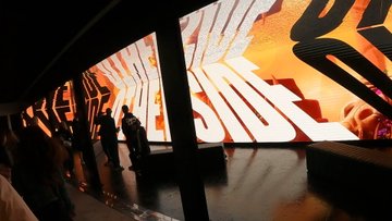 A dimly lit room with curved walls displaying bright orange digital projections, where silhouetted figures stand watching the immersive "Bored Ape Yacht Club" exhibition installation.
