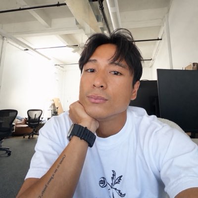 A close-up selfie of an Asian man wearing a white t-shirt, resting his chin on his hand, with an office environment visible in the background.