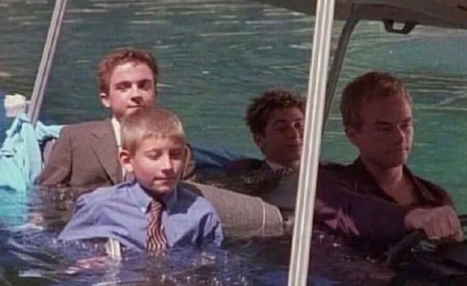 Four men sit calmly in a car that has sunk deep into water, with water reaching up to their chests, as they appear unfazed by the flooded situation. The image humorously relates to the post by depicting the guys' stoic composure despite being in a dire situation, mirroring the idea of holding steady and not "selling" on each other while the group chat cook faces backlash.