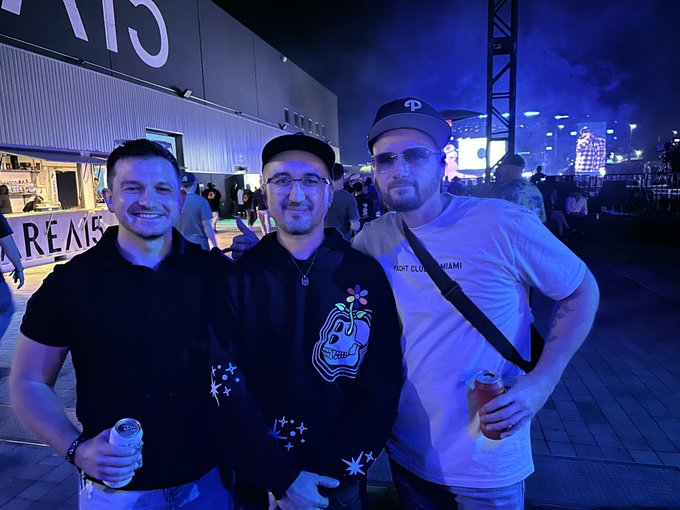 Three men posing together at an outdoor night event, with one holding a canned drink and another wearing a cap marked with a 'P' logo.