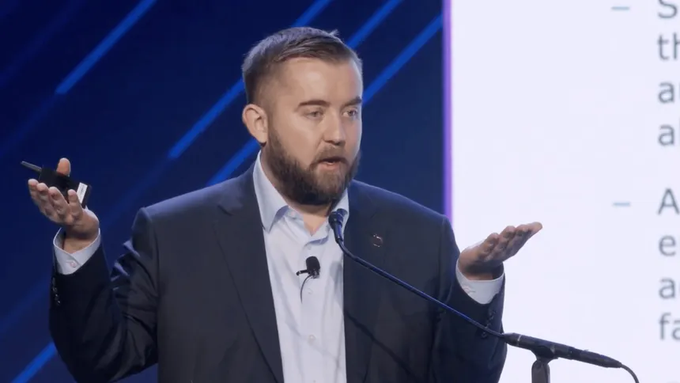 A bearded man in a dark suit and light shirt speaks expressively at a podium with both hands raised, standing in front of a presentation screen.