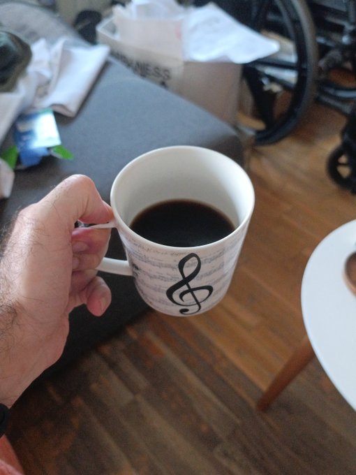 A hand holds a white coffee mug decorated with a treble clef and musical notes, filled with black coffee. In the background, a wheelchair, a Hermes box, and various items are visible on a dark surface next to a wooden floor.