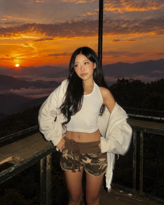 A young woman with long dark hair poses for a photo wearing a white crop top, camo shorts, and a white jacket, set against a scenic sunset over mountains.