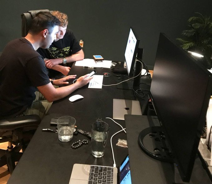 Two young men sitting at a dark desk in an office setting, looking at a smartphone while working on a laptop with a large monitor nearby, with water glasses, a MacBook, and various items scattered on the desk.