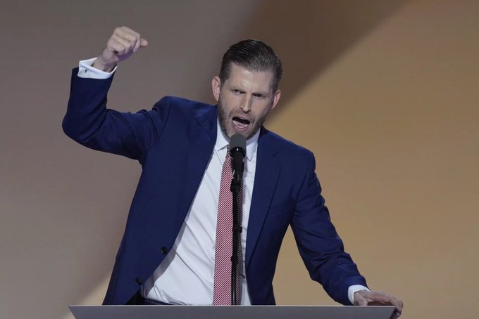 Eric Trump in a blue suit and red tie raises his fist passionately at a podium with a microphone, likely at the 2024 RNC.