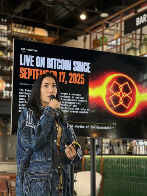 A woman with long dark hair wearing a denim jacket speaks into a microphone in front of a banner reading 'LIVE ON BITCOIN SINCE SEPTEMBER 17, 2025' featuring a Bitcoin logo design.