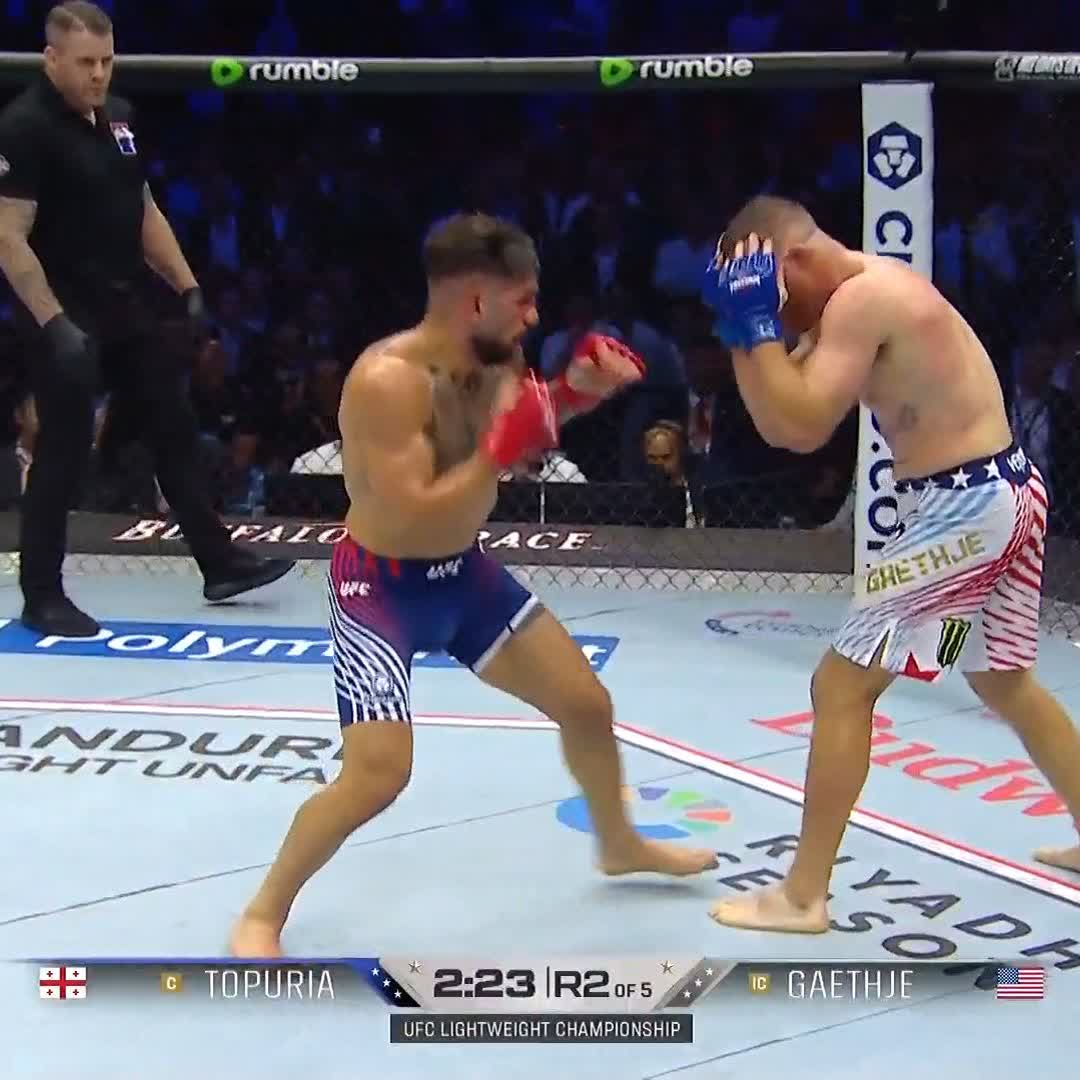 The image shows a fight between two boxers, with one boxer wearing blue shorts and the other wearing red shorts. The fighter in blue shorts appears to be dominating his opponent as he throws a punch towards him. The fighter in red shorts is caught off guard by this move and is struggling to defend himself. The post highlights that the fight is taking place at the UFC Lighthouse, with the fighters being referred to as "@TopuriaIlia" and "@Cryptocom".