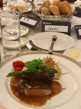A plated meal featuring meat, garnishes, and sauce, served on a white plate at a dining table set with glasses of water and bread, with a 'Crobi' name card visible.
