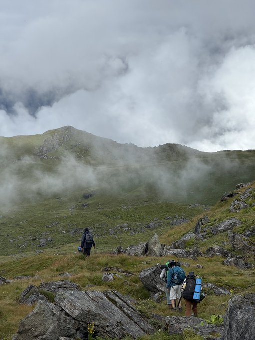 Two hikers with backpacks traverse a grassy, rocky mountain slope as mist rolls over the ridge above them.