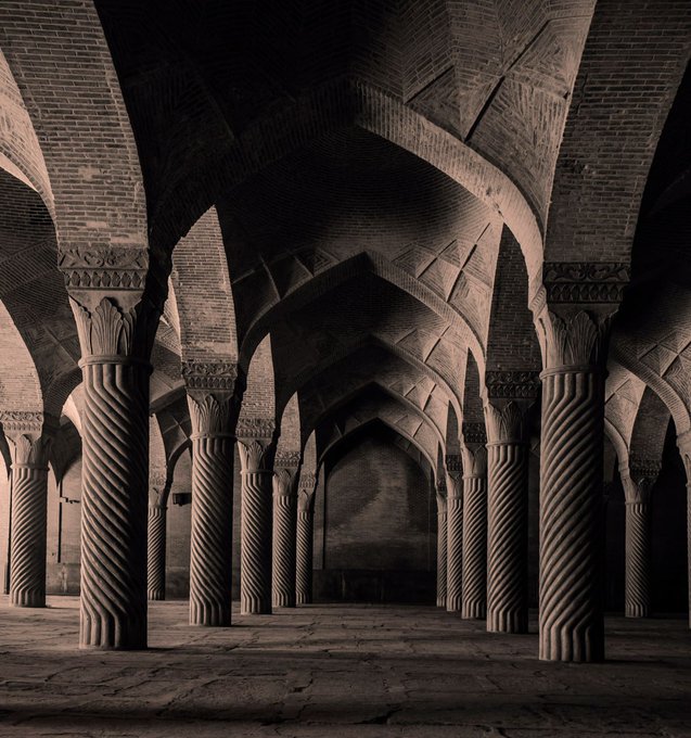 An atmospheric view of a historic Persian-style mosque interior with intricately carved brick arches and spiral-fluted columns, cast in deep shadows.