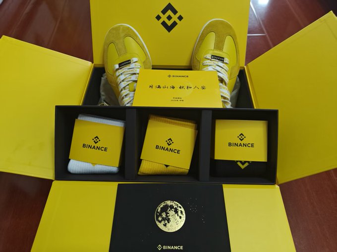 An open yellow Binance promotional gift box containing a pair of yellow sneakers, white socks, and yellow Binance-branded boxes. A booklet with Chinese text and a gold-colored Binance-branded moon coin sit in the foreground.