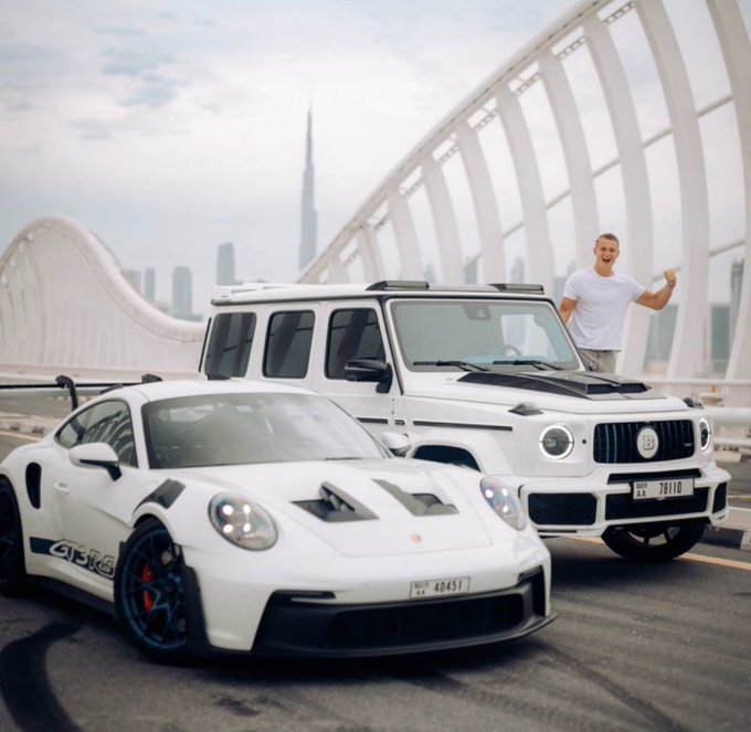 A man poses between a white Porsche 911 GT2 RS (license plate 40451) and a white Mercedes-Benz G-Wagon (license plate 78110) on a Dubai overpass with the Burj Khalifa in the background, symbolizing the lavish crypto trading lifestyle described in the caption as the reward for surviving multiple Bitcoin market cycles.