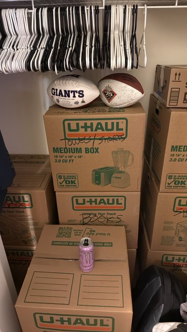 U-Haul moving boxes are stacked in a closet, topped with signed New York Giants and 49ers footballs, with hangers full of clothes hanging above and a pink Monster Energy drink can on top.
