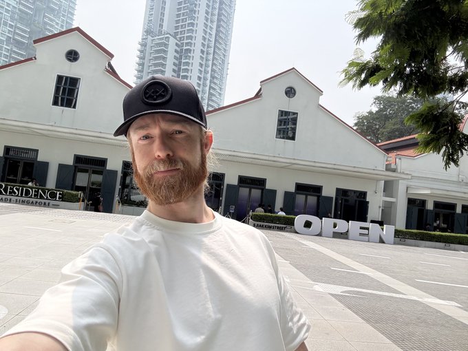 A selfie of a bearded man wearing a black baseball cap with a logo and a white shirt, taken outside a building marked 'RESIDENCE' and 'OPEN' with modern high-rise buildings in the background.