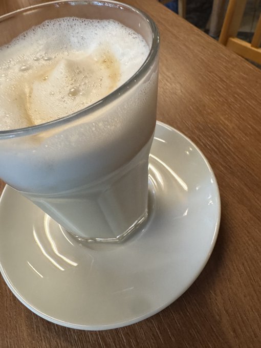 A close-up of a frothy coffee drink in a glass served on a white saucer atop a wooden table.