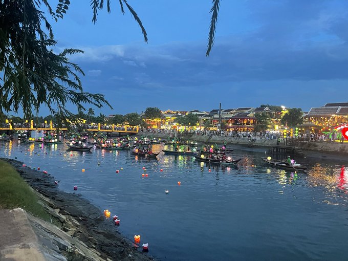 A scenic view of a river at dusk in Hoi An, Vietnam, with boats floating on the water, lanterns lining the riverbank, and an illuminated bridge in the background.