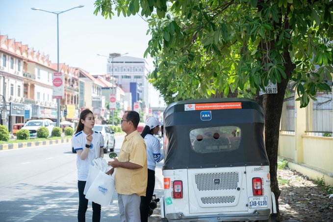 Two ABA bank employees in uniform converse with a tuk-tuk driver beside his vehicle on a sunny street in Phnom Penh, illustrating post's theme of real-world vehicle lease activity that underpins Musubi A/R's on-chain yield.

License plate "1AB-6198" and tiny "55 58 77" sticker are visible on the tuk-tuk, along with a red "Cambodia" banner across the windshield.