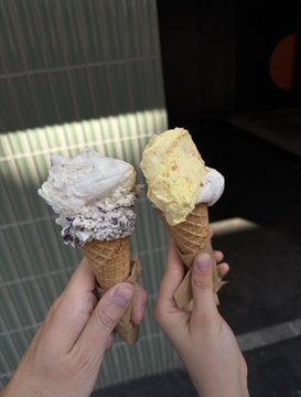 Two hands holding ice cream cones, one with cookies and cream flavor and the other with a yellow flavor.