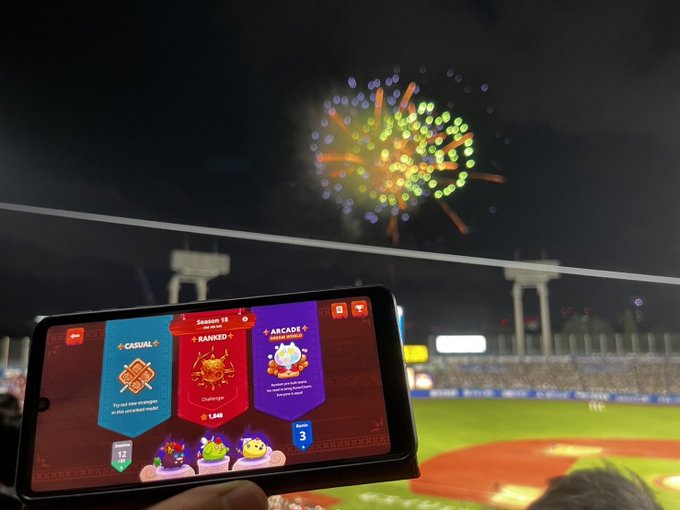 A phone displaying a stadium game's reward screen is held up against a night sky with fireworks.