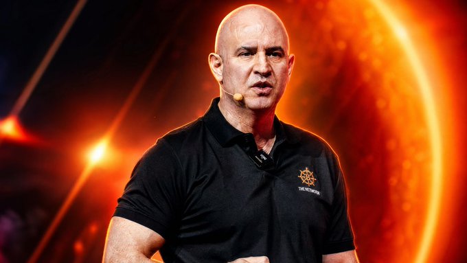 The image features a bald man wearing a black shirt, standing in front of a large orange circle with a microphone attached to his face. The background is dark and blurred, drawing attention to the main subject - Tony Yazbeck, who appears to be delivering a keynote speech at an event.