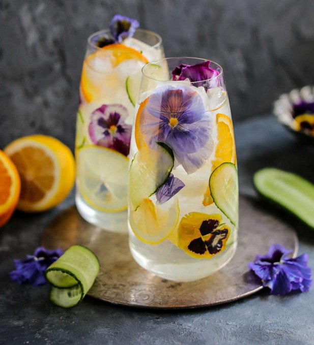 Two tall glasses of infused water garnished with orange slices, cucumber, lemon, and purple pansies on a dark surface.