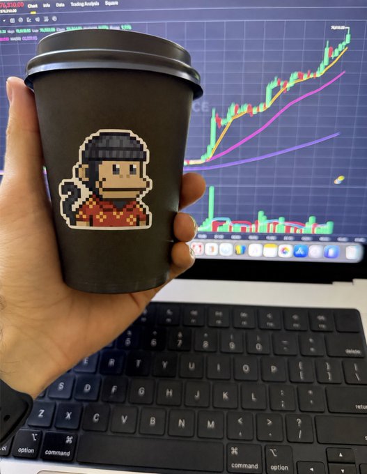 A hand holding a black coffee cup featuring a pixel art monkey character in a beanie, with a laptop displaying a stock trading chart showing green and red candlesticks in the background.