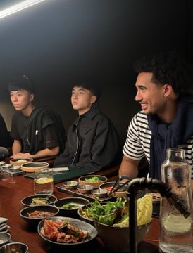 Three young men sit at a restaurant table with plates of Korean food and various side dishes (banchan) spread before them, with one man on the right smiling at the others.