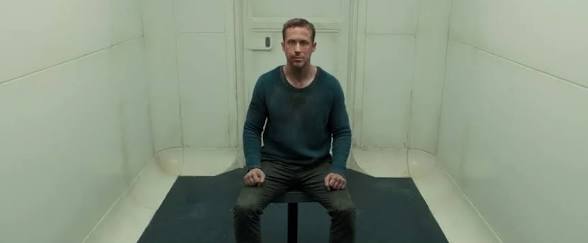 A man sits alone on a stool in a stark, white, padded room, looking directly at the camera.