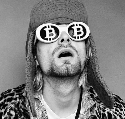 A black-and-white photo shows a man with long hair and a goatee, wearing a furry hat and distinctive round sunglasses featuring the Bitcoin "₿" logo, his mouth open in an expressive pose. The Bitcoin-themed eyewear visually ties into the post's discussion of crypto developments, symbolizing the digital currency ethos central to the Base (Coinbase's L2) leadership transition being announced.
